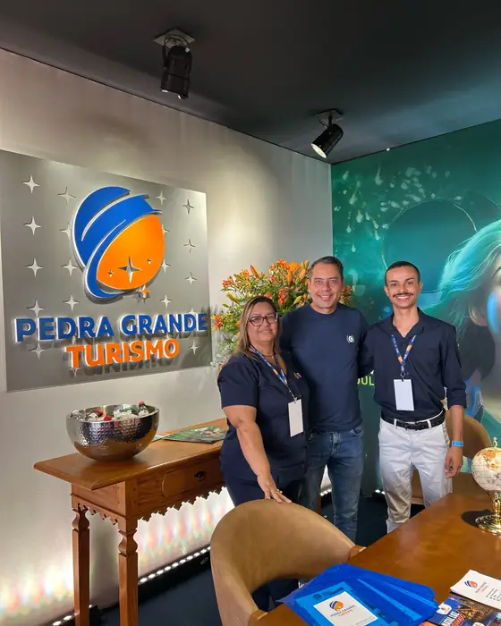 Três pessoas da equipe com crachá, ao lado do logotipo da Pedra Grande iluminado na parede do estande.